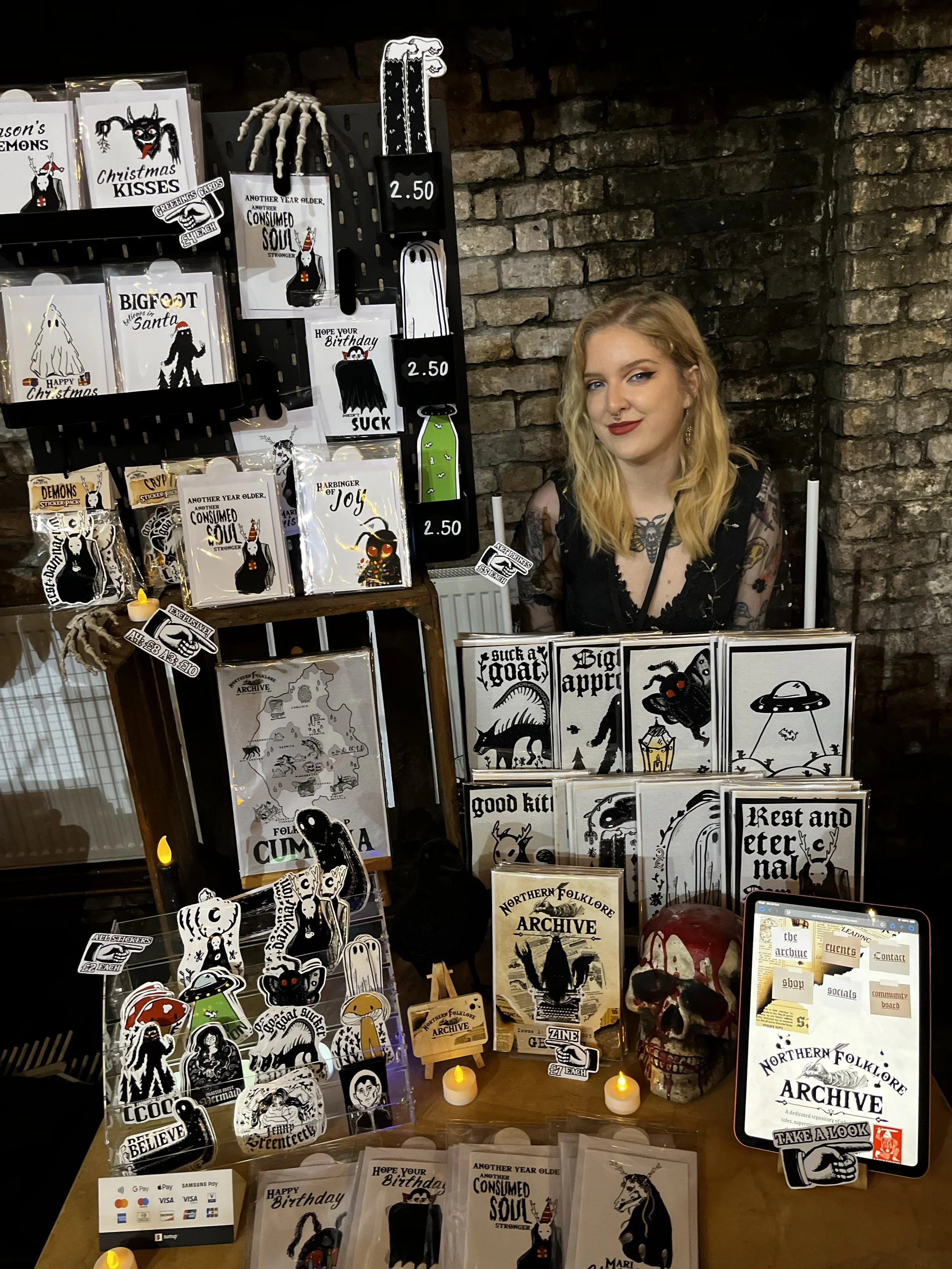 A woman with blonde hair and red lipstick behind a display of black and white folklore-themed merchandise, including cards, stickers, and a skull, at a market stall against a brick wall.