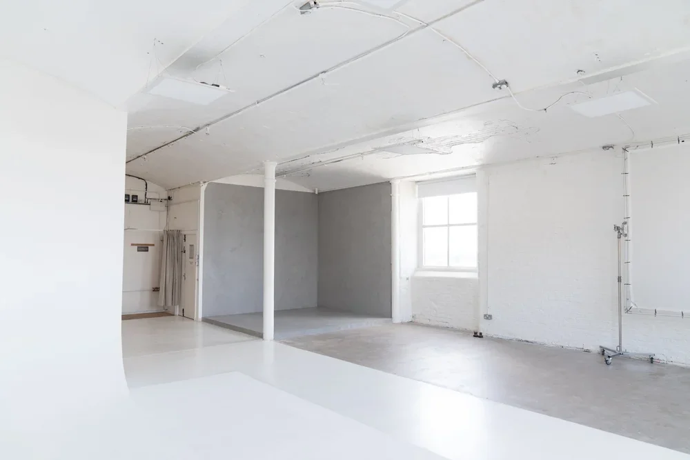 Empty room with white walls, exposed ceiling pipes, a large window, and a section with gray walls and flooring.
