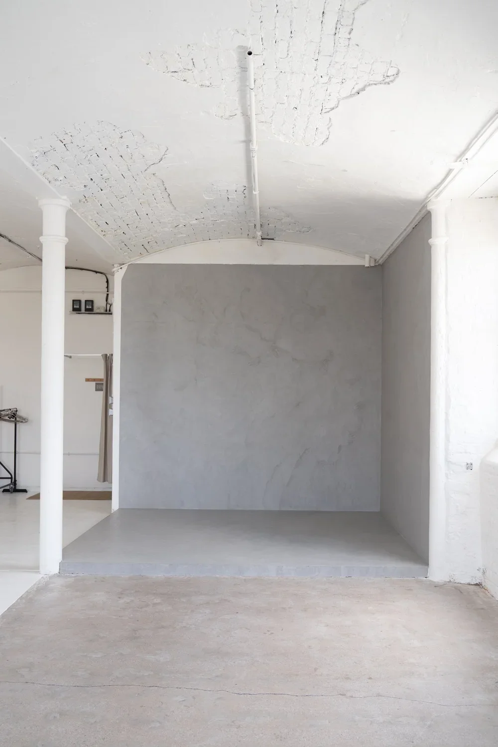 Minimalist white interior space with a small raised platform, white brick ceiling with some exposed brick patches, and a column on the left. There's a doorway with a curtain on the left side.