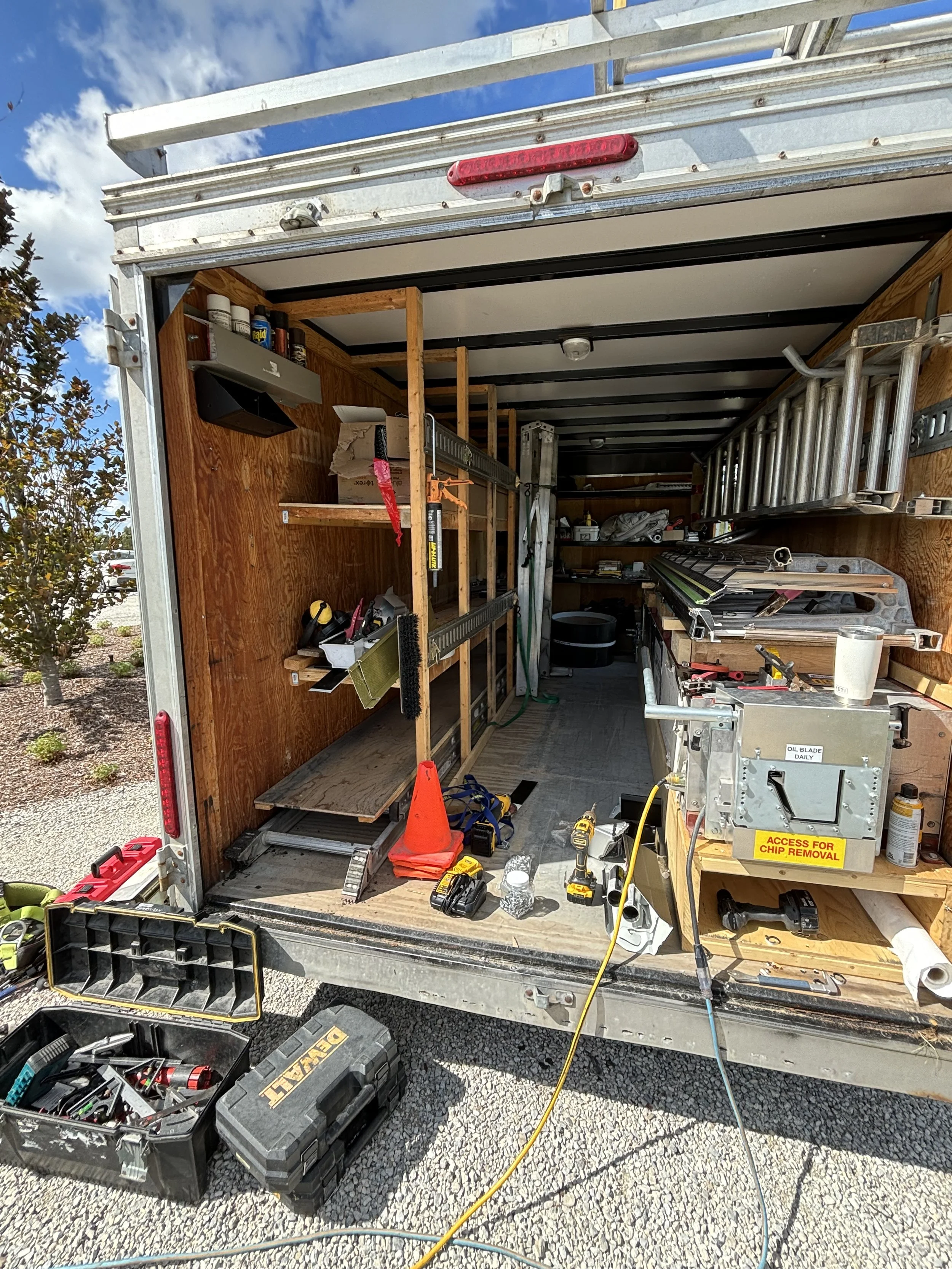 Interior of a mobile workshop or trailer with tools and equipment, including ladders, power drills, a level, and shelves with supplies, set up outdoors on a gravel surface.