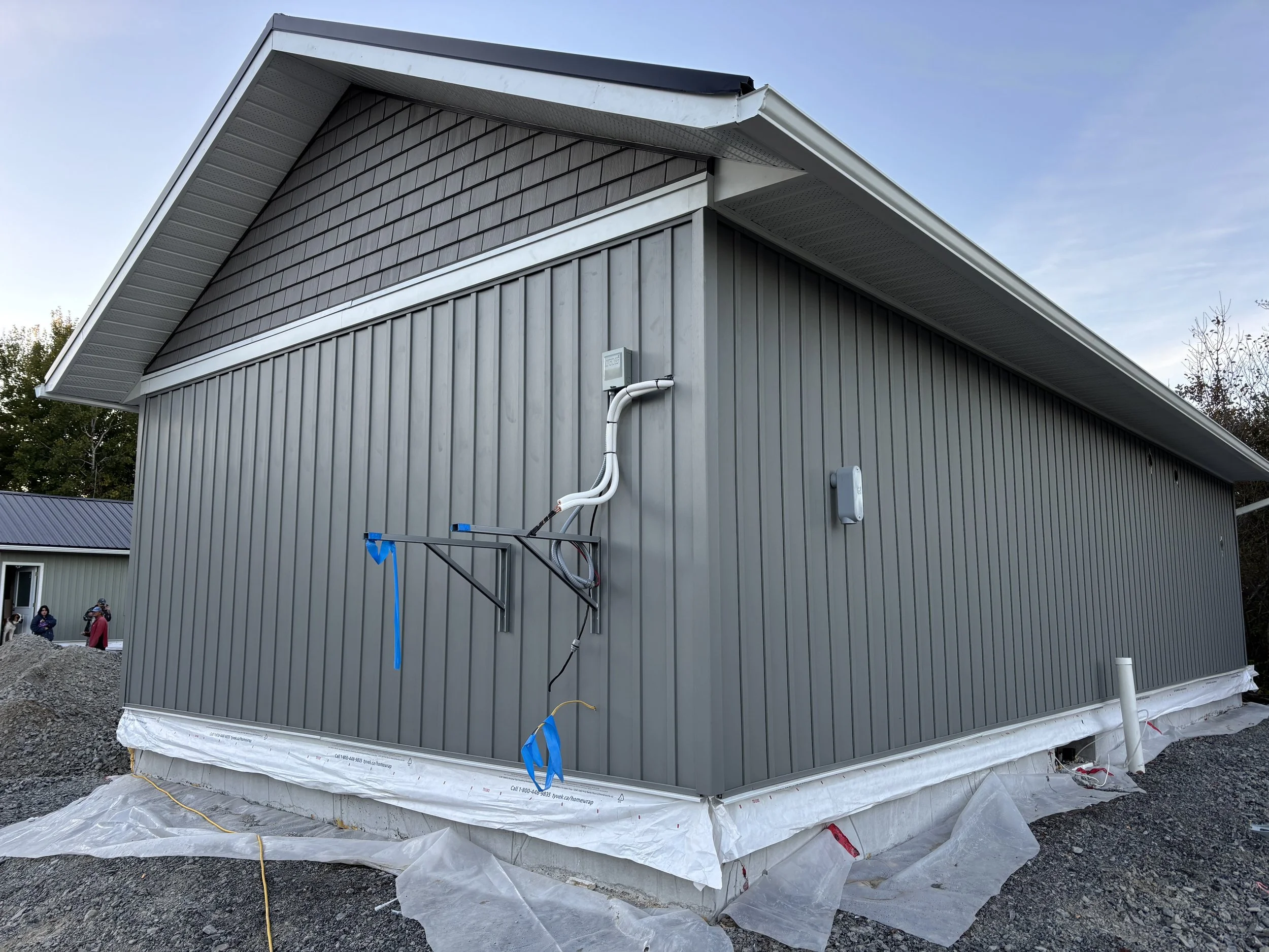 Exterior of a gray building under construction with electrical wiring and conduit on its side, with workers and a small building visible in the background.