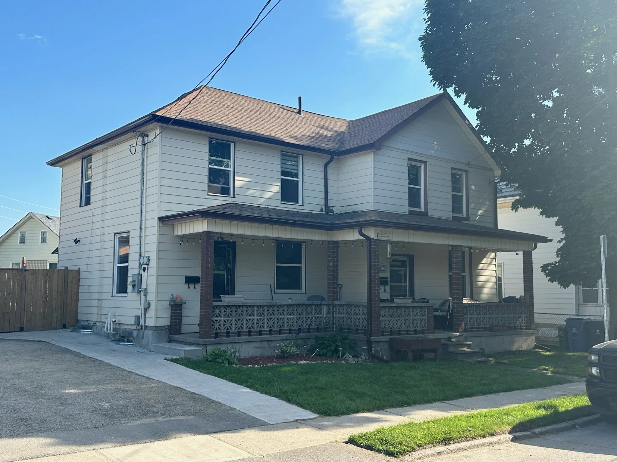 A two-story white house with a front porch, brick supports, and a small lawn, situated in a suburban neighborhood under a clear blue sky.