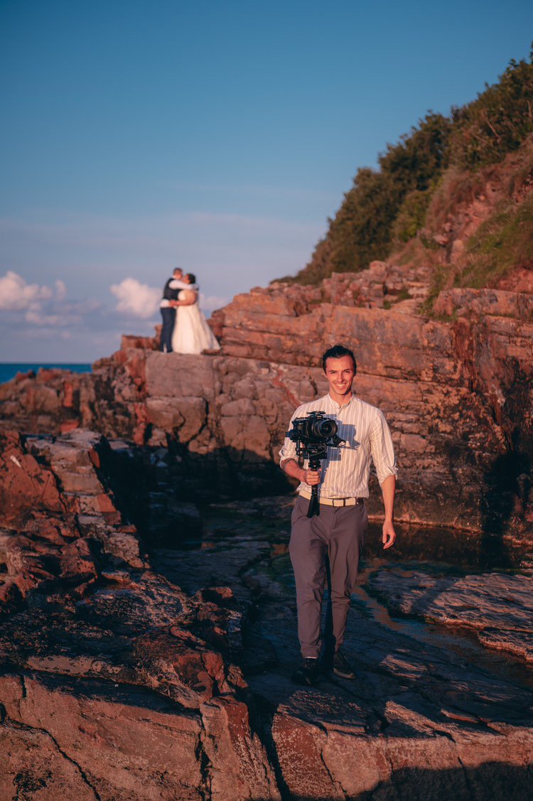 A photographer standing on rocks near the water, capturing a couple embracing on a rocky shoreline with greenery on a hillside in the background during sunset.