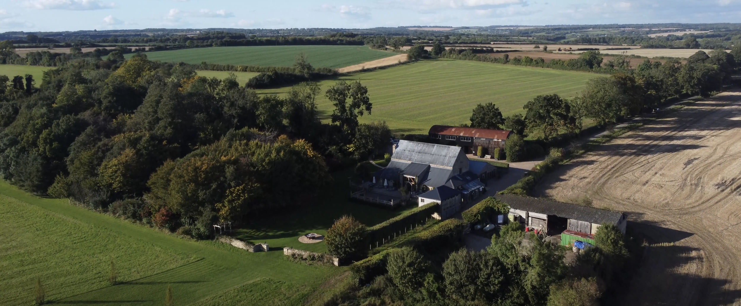Aerial view of a rural landscape with fields, trees, and farm buildings, including a house, barn, and storage structures. Cripps Barn