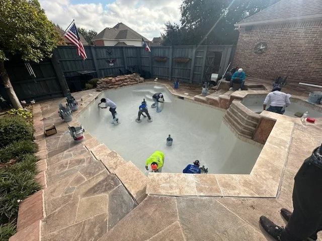 Workers are renovating a residential backyard swimming pool with a stone border, using power tools and equipment. The pool is empty, and workers are inside and around it. An American flag flies in the background near a wooden fence, and a brick house is adjacent. The sky is partly cloudy.