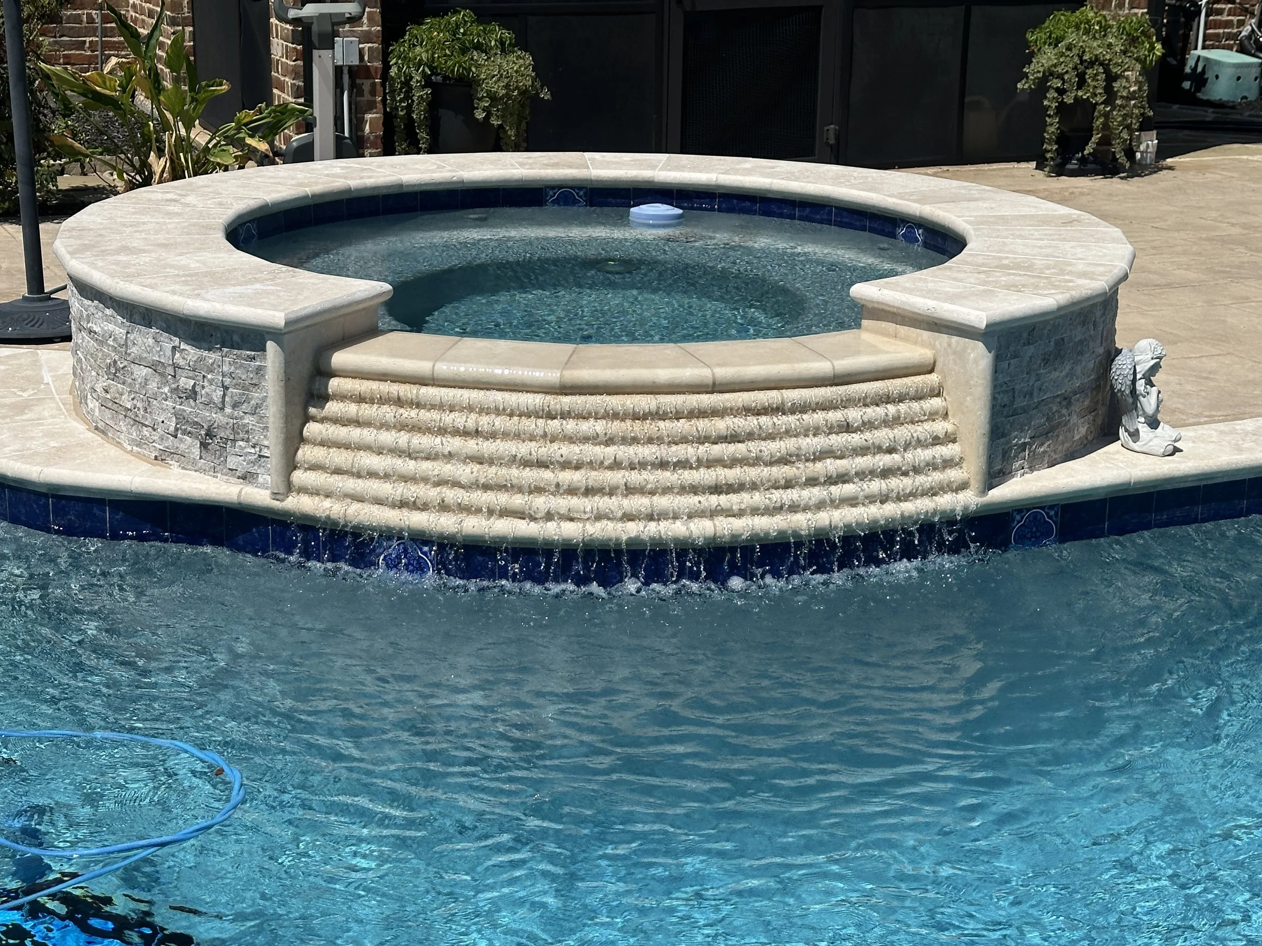 A newly remodeled backyard hot tub with stone and tile construction next to a swimming pool, decorated with small statues and surrounded by patio furniture and potted plants.