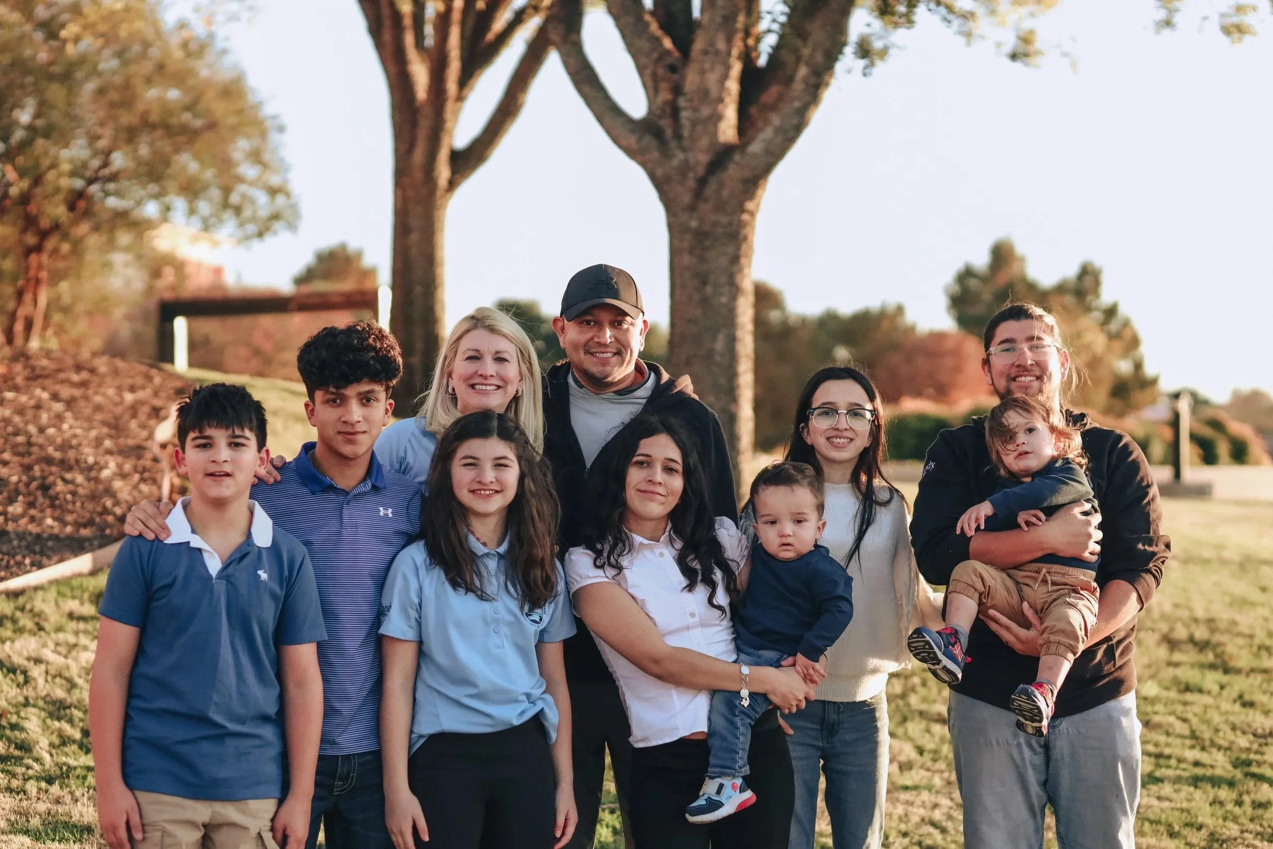 A family of ten people standing outdoors in front of trees, smiling, with a sunny sky.