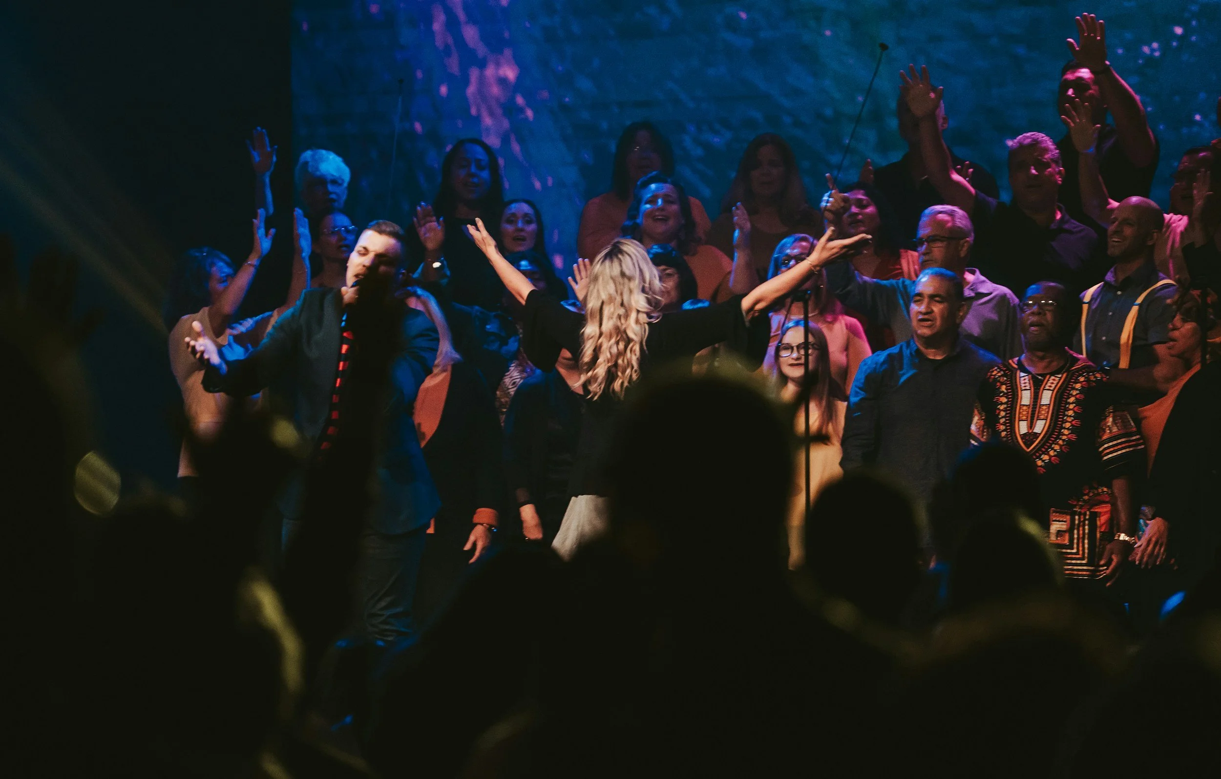 People singing and worshiping with their hands raised on stage during a concert or religious gathering, with a singer in front and a screen with abstract visuals in the background.