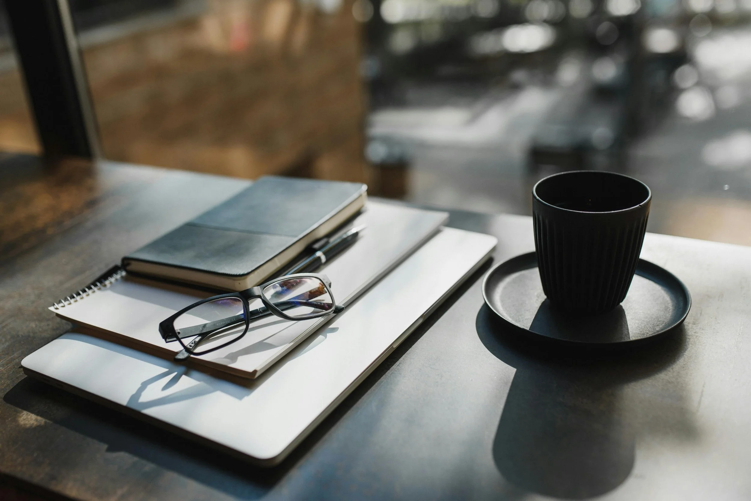 A black coffee cup on a saucer, placed on a table. Next to it are notebooks, a pen, and eyeglasses on top of a closed laptop. Sunlight creates shadows on the table.