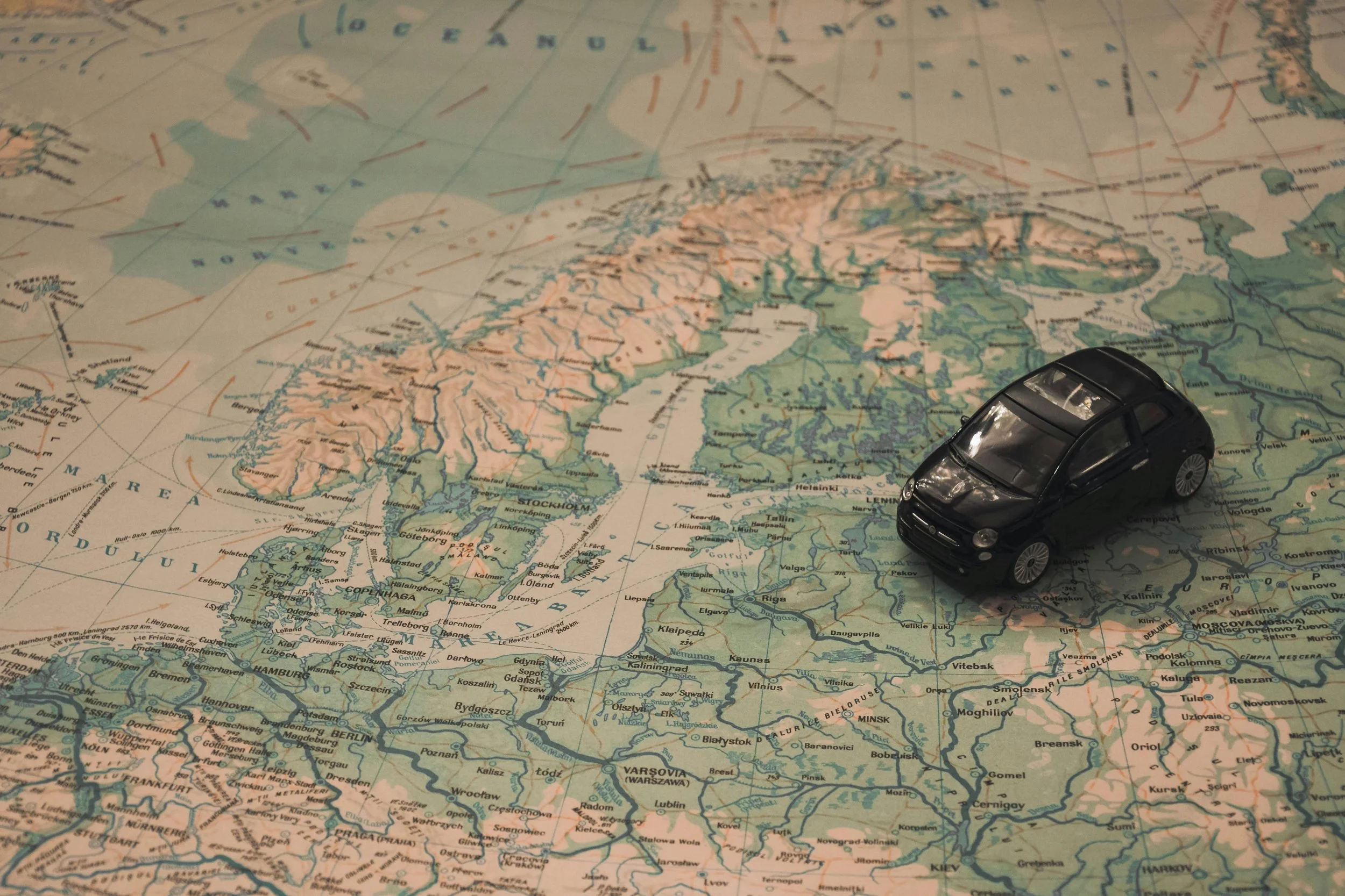 A small, black toy car drives across a sepia and green map of northern Europe.