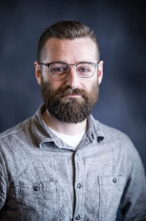 A man with glasses, a full beard, and short hair, wearing a gray button-up shirt, looking at the camera with a slight smile against a dark background.
