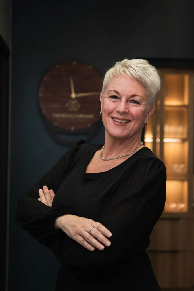 An older woman with short, white hair smiling, wearing a black top and a silver necklace, standing with arms crossed in front of a dark wall with a round wooden clock in the background.
