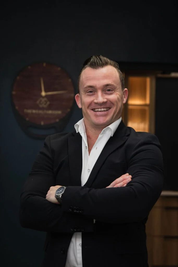 Smiling man in a black suit with crossed arms, standing in front of a dark background with a round clock on the wall.