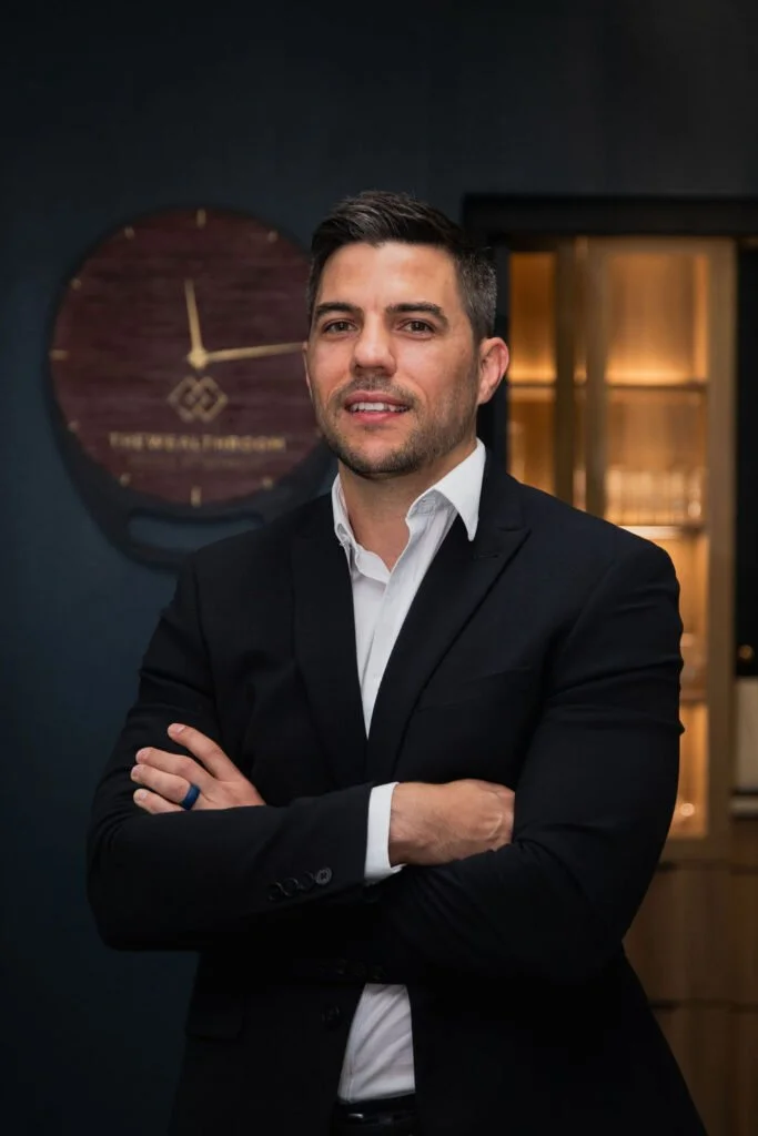 A man in a black suit with a white shirt standing with arms crossed, in an office setting with a large wall clock behind him.