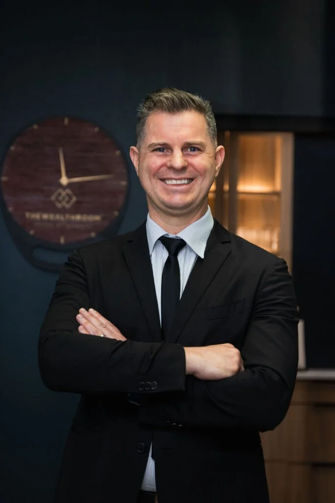 A smiling man in a black suit with a light blue dress shirt and black tie standing with arms crossed, in an office setting with a clock on the wall behind him.