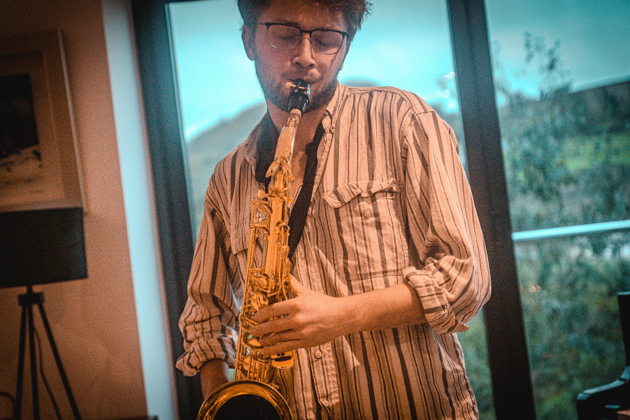 A young man with glasses wearing a striped shirt, playing a saxophone indoors with large windows and trees visible outside.