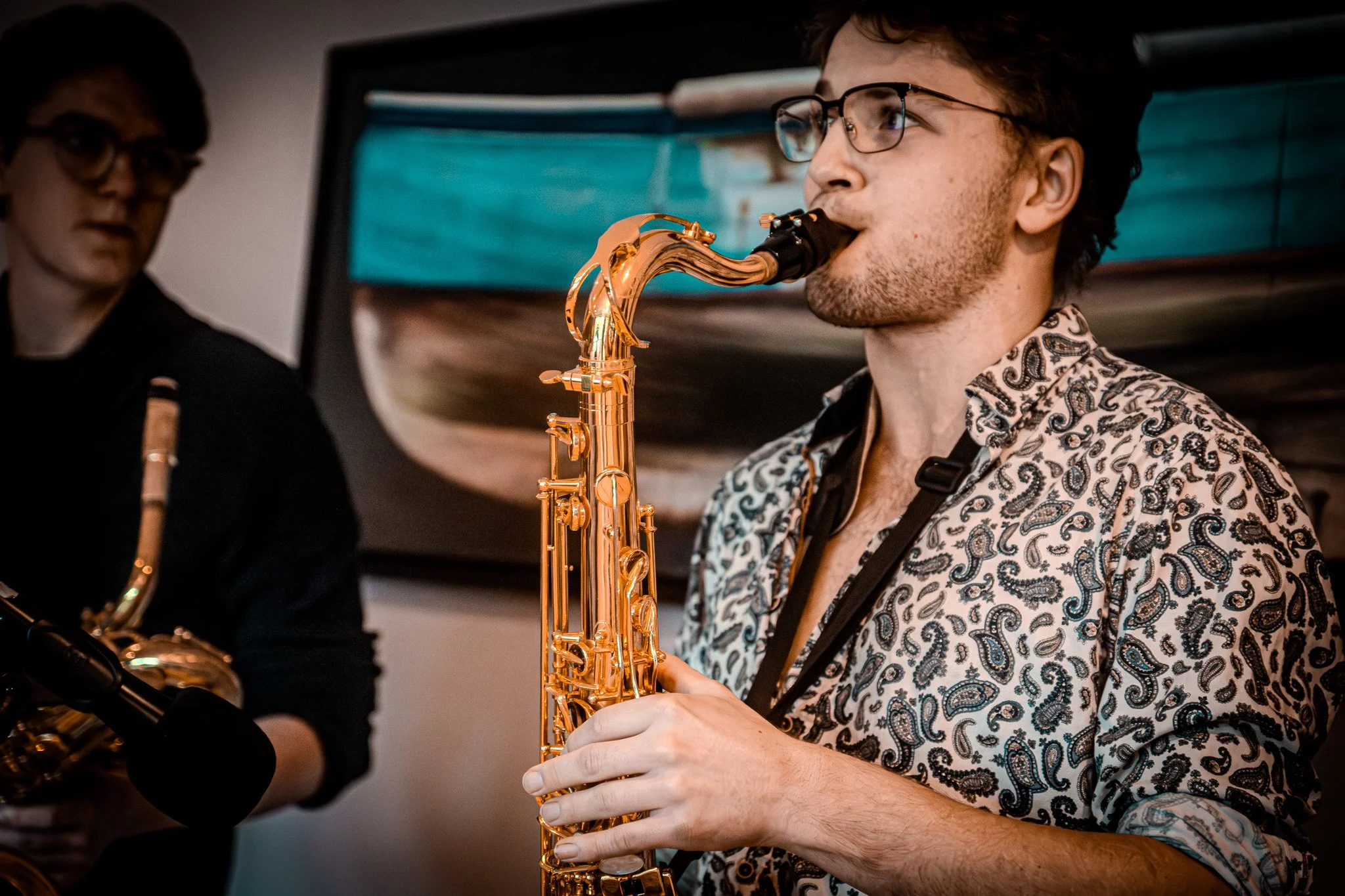 A young man with glasses playing a gold saxophone, wearing a patterned shirt. Another person with glasses holding a saxophone stands nearby, with a painting of a boat in the background.
