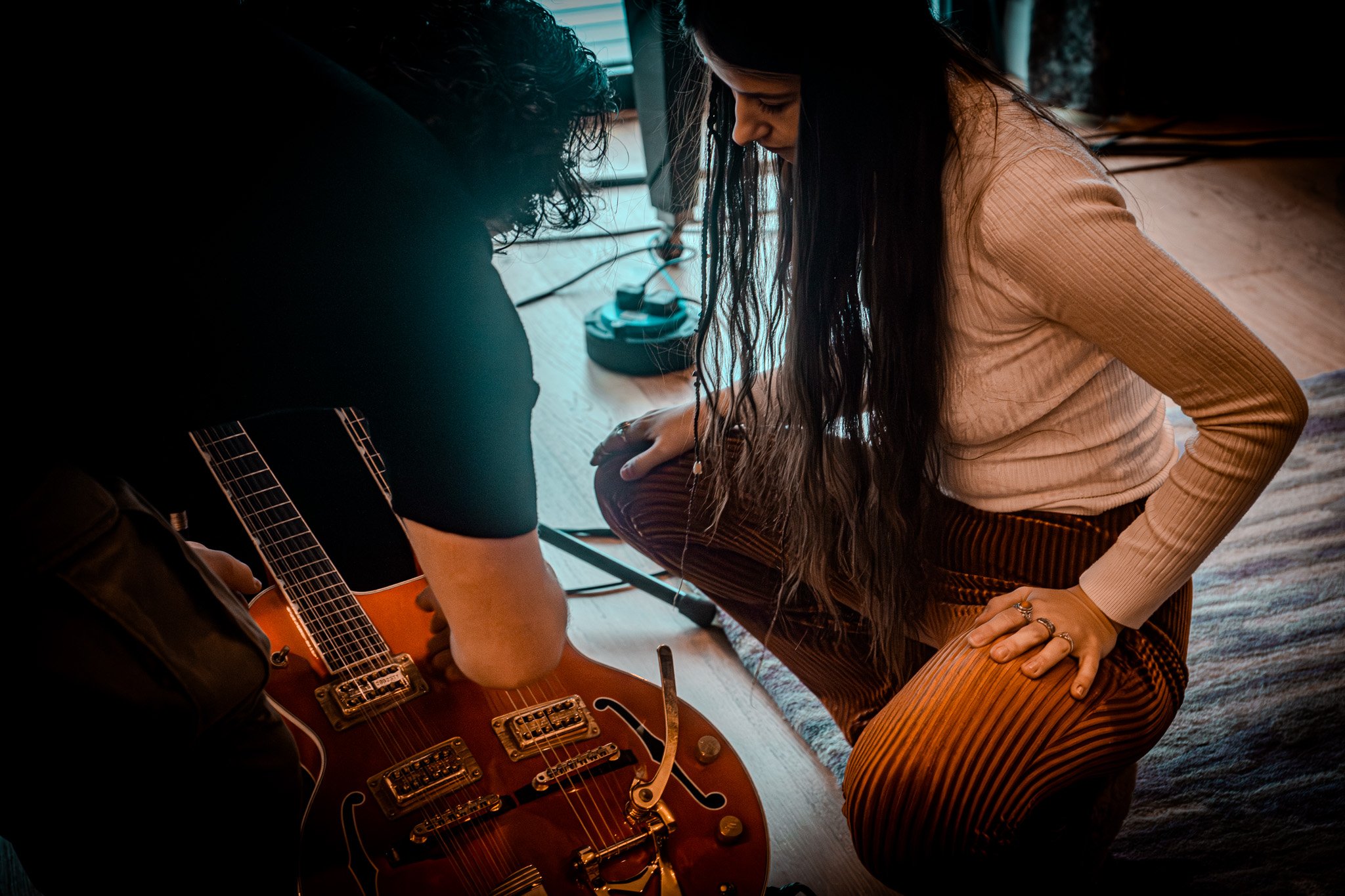 Two women kneeling on a wooden floor, one with long dark hair wearing a beige sweater and striped pants, the other with curly hair in a black shirt, tuning a orange electric guitar.