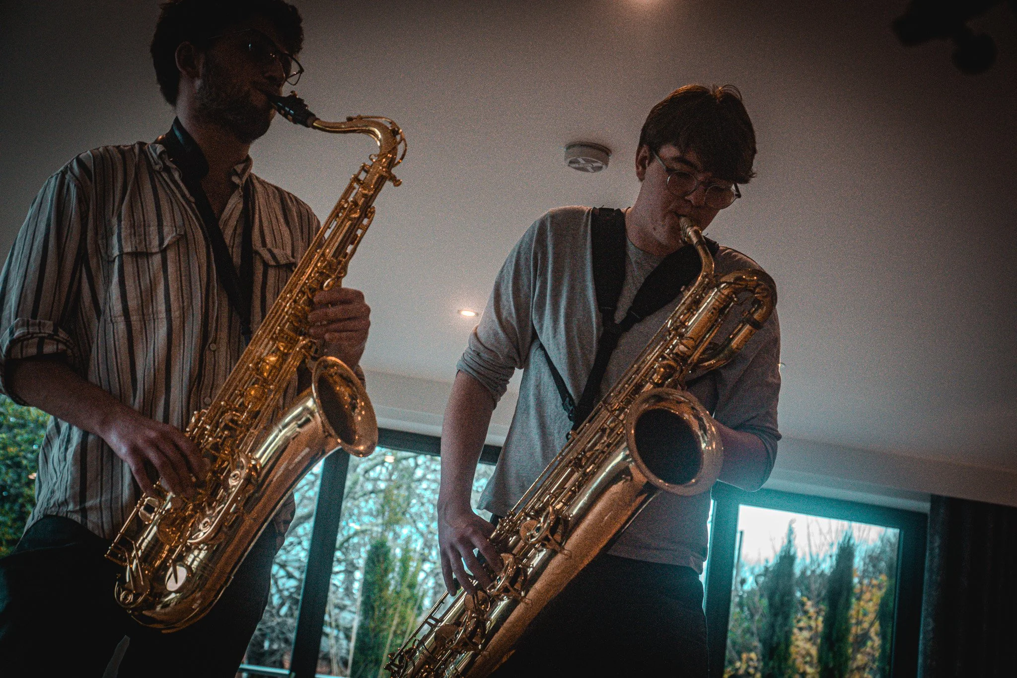 Two men playing saxophones indoors with large windows in the background revealing trees outside.