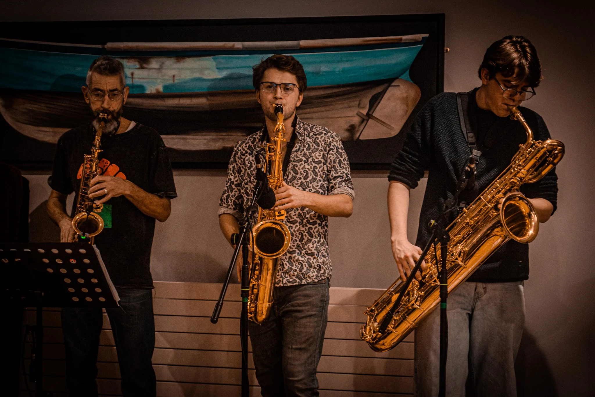 Three musicians playing saxophones during a live performance, with a painting of a boat behind them.