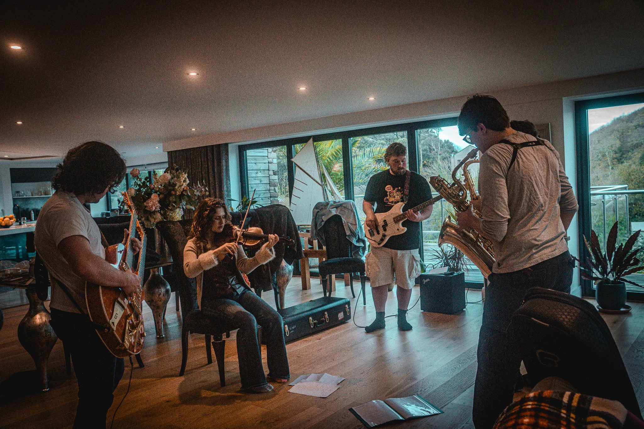 A group of five musicians playing instruments in a living room, with large windows showing trees outside.