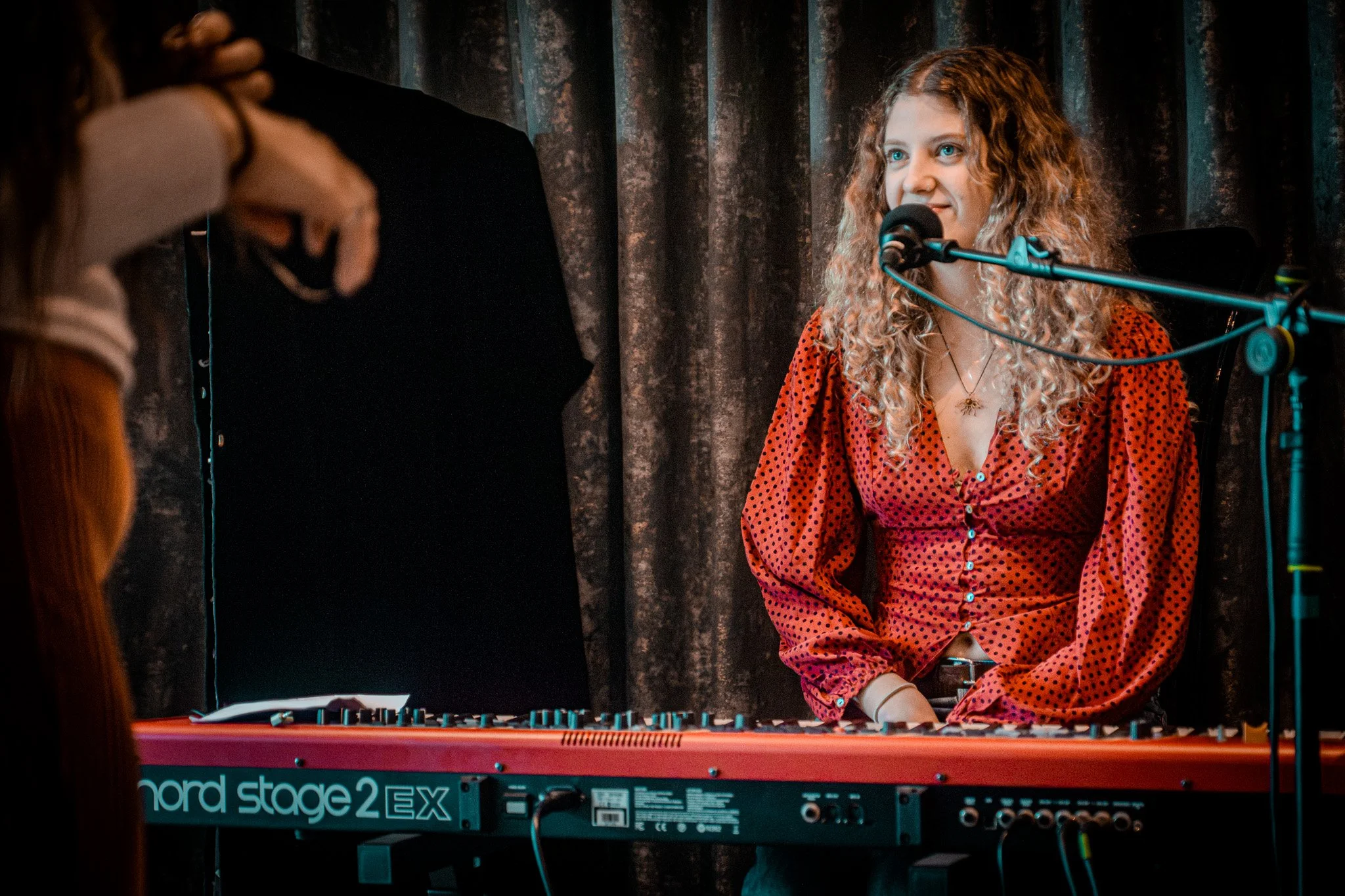 A young woman with curly blonde hair singing into a microphone behind a keyboard on stage, with a person in the foreground, against a dark curtain background.