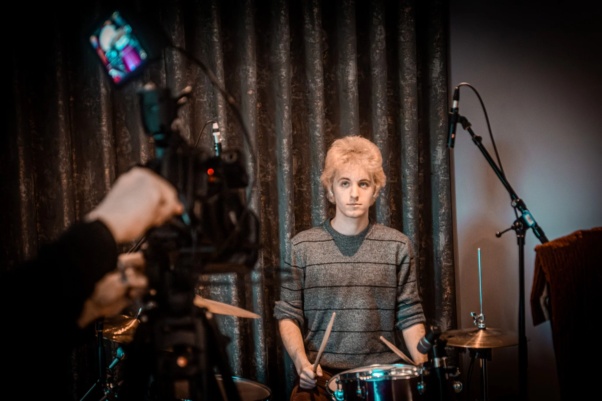 A woman with short blonde hair stands behind a drum set, looking towards a camera. The scene appears to be in a recording studio with dark curtains in the background.