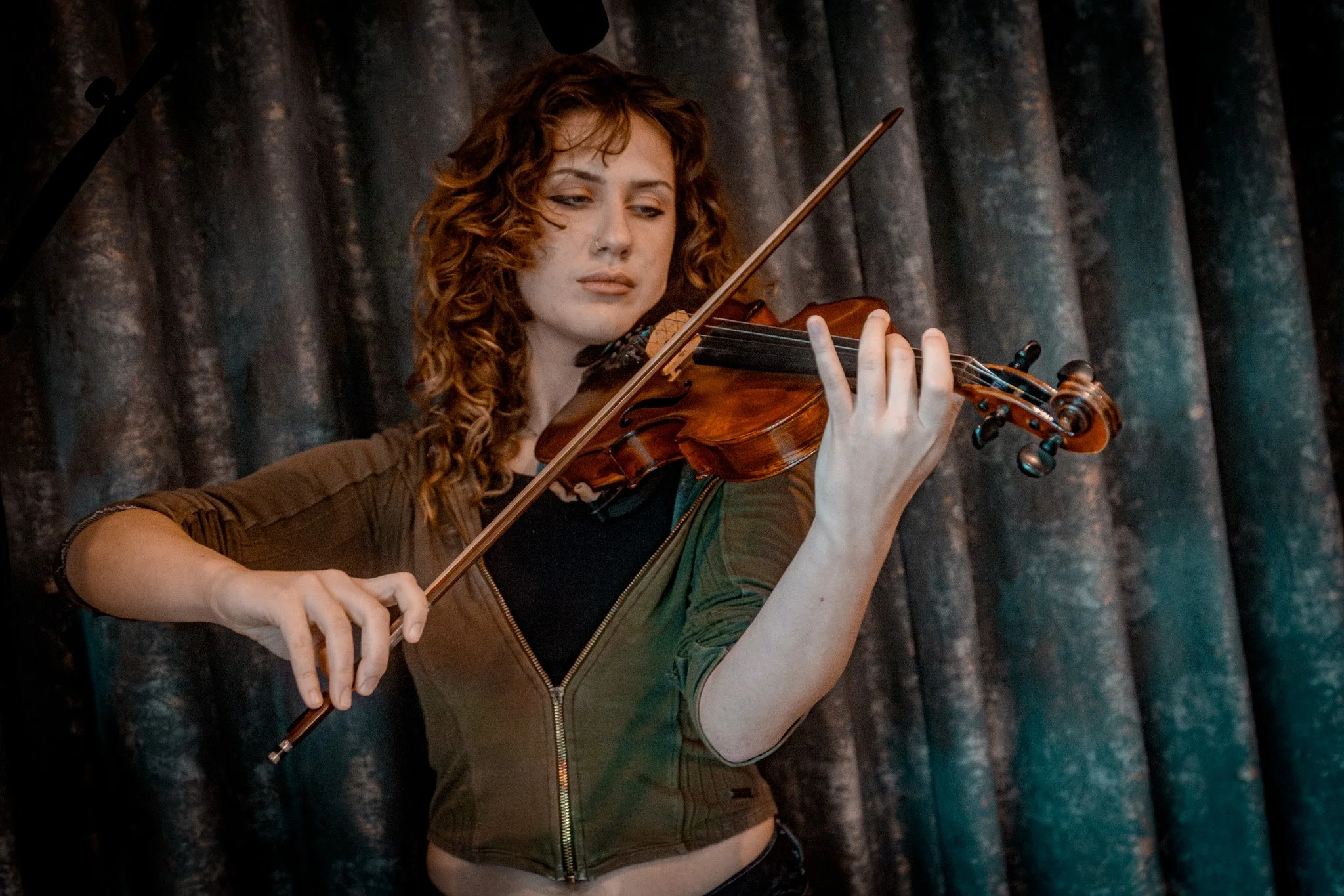 A young woman with curly hair playing a violin on stage in front of a dark curtain.