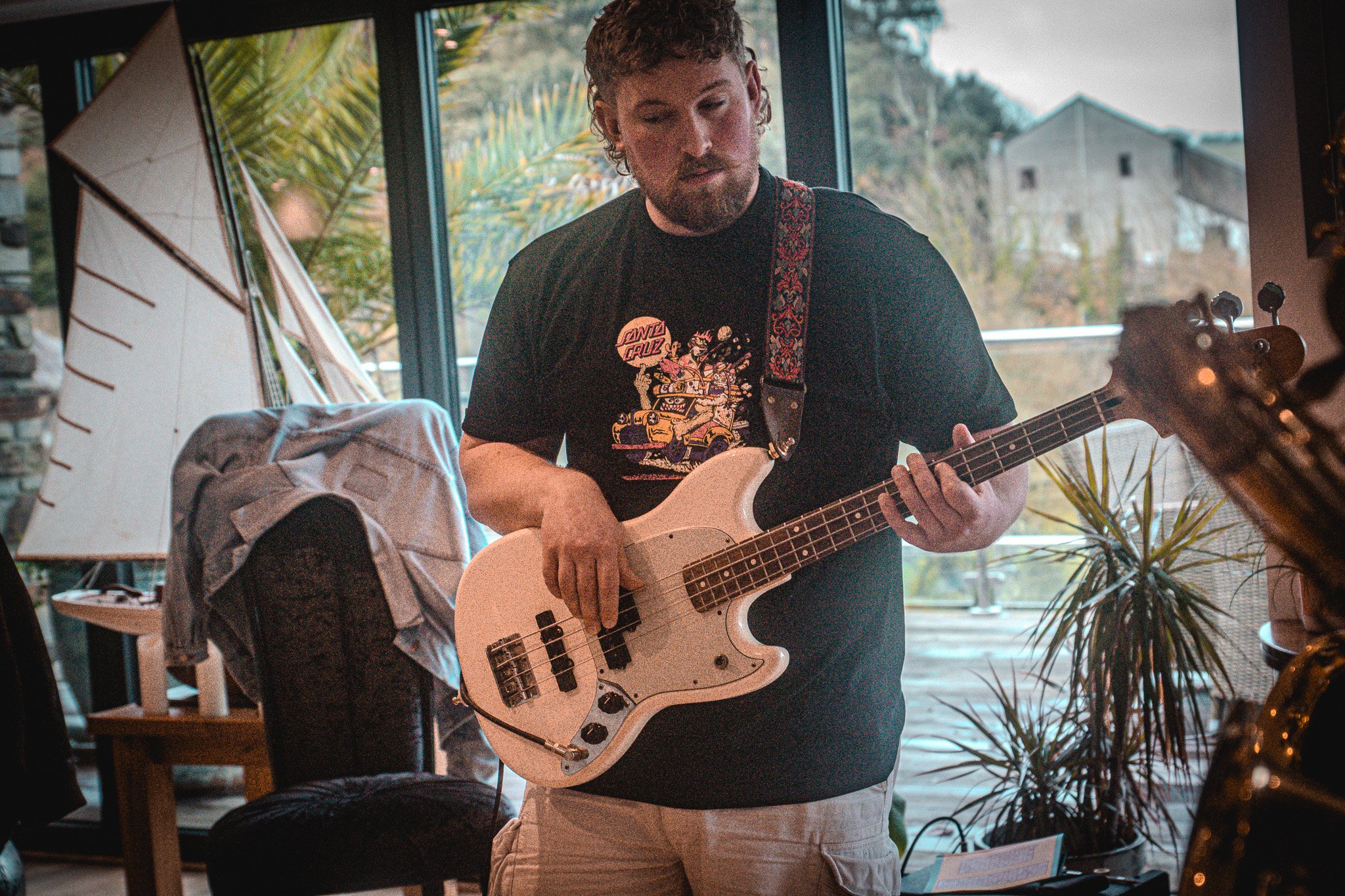 A man playing an electric bass guitar indoors with large windows and plants in the background.
