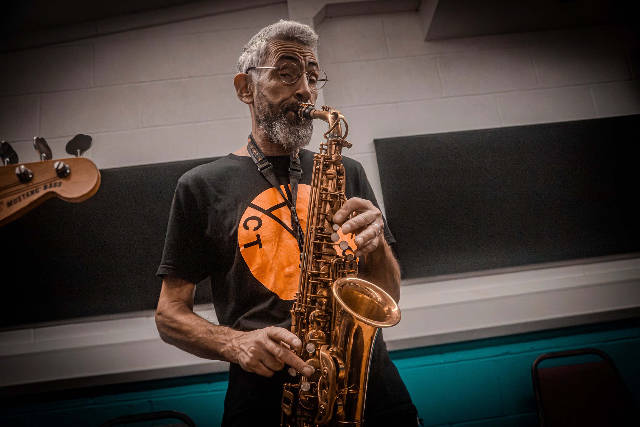 A man with glasses, gray hair, and a beard playing a saxophone in an indoor setting.