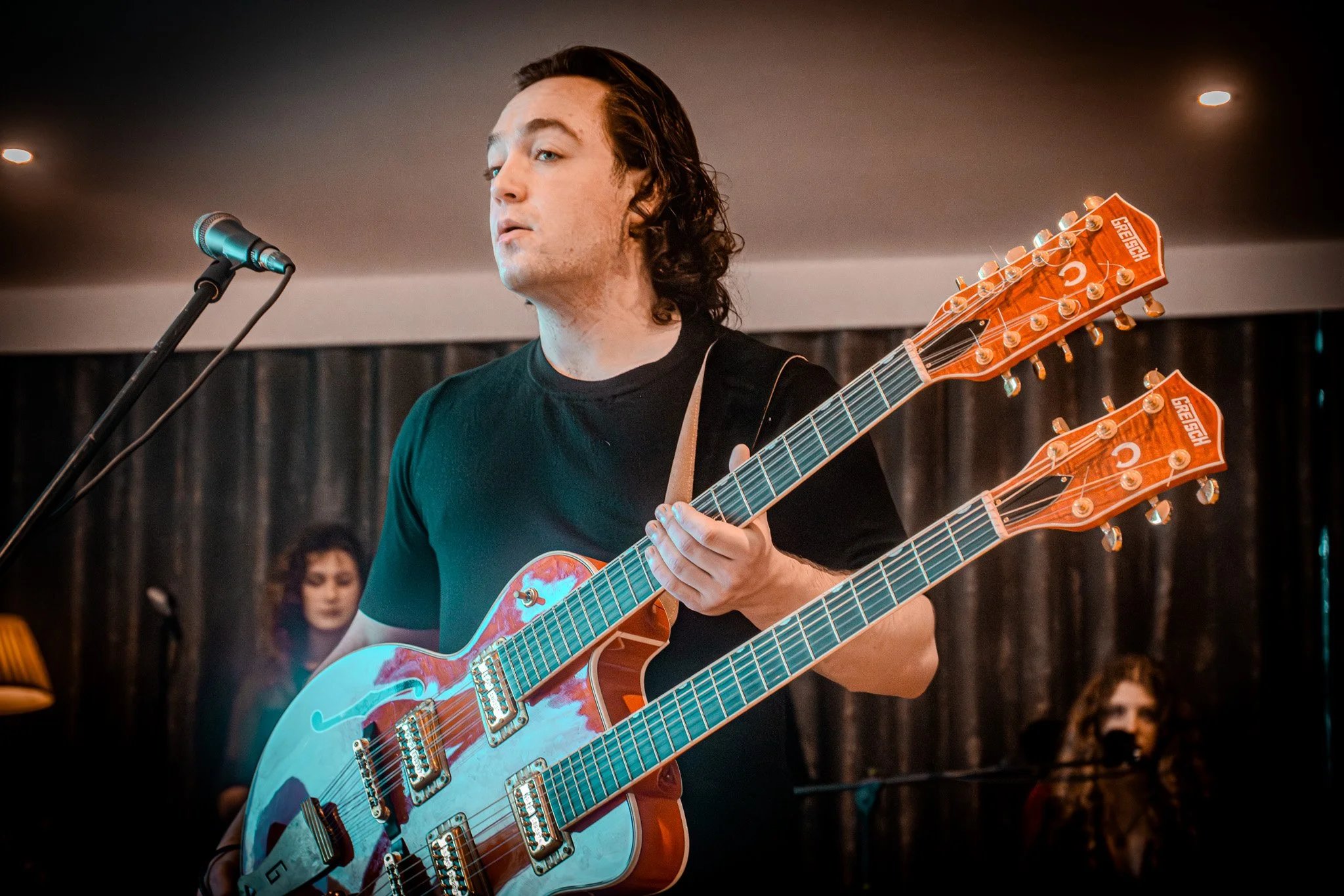 A musician performing with two double-neck electric guitars with orange wood finish, wearing a black shirt, standing in front of a microphone, with a woman and a person in the background.