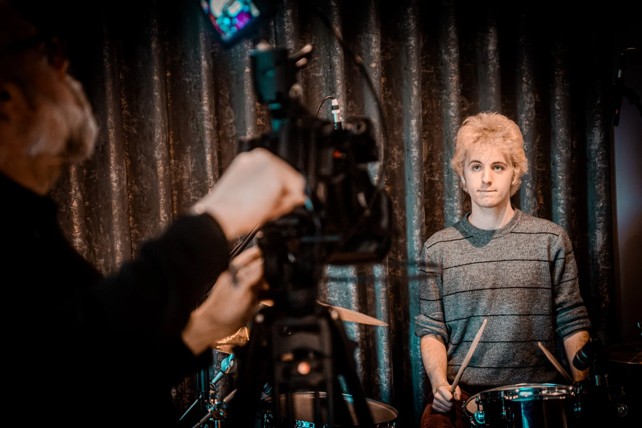 A young man with curly blonde hair and a gray striped sweater sitting at a drum set, being filmed or photographed by a person operating a camera.