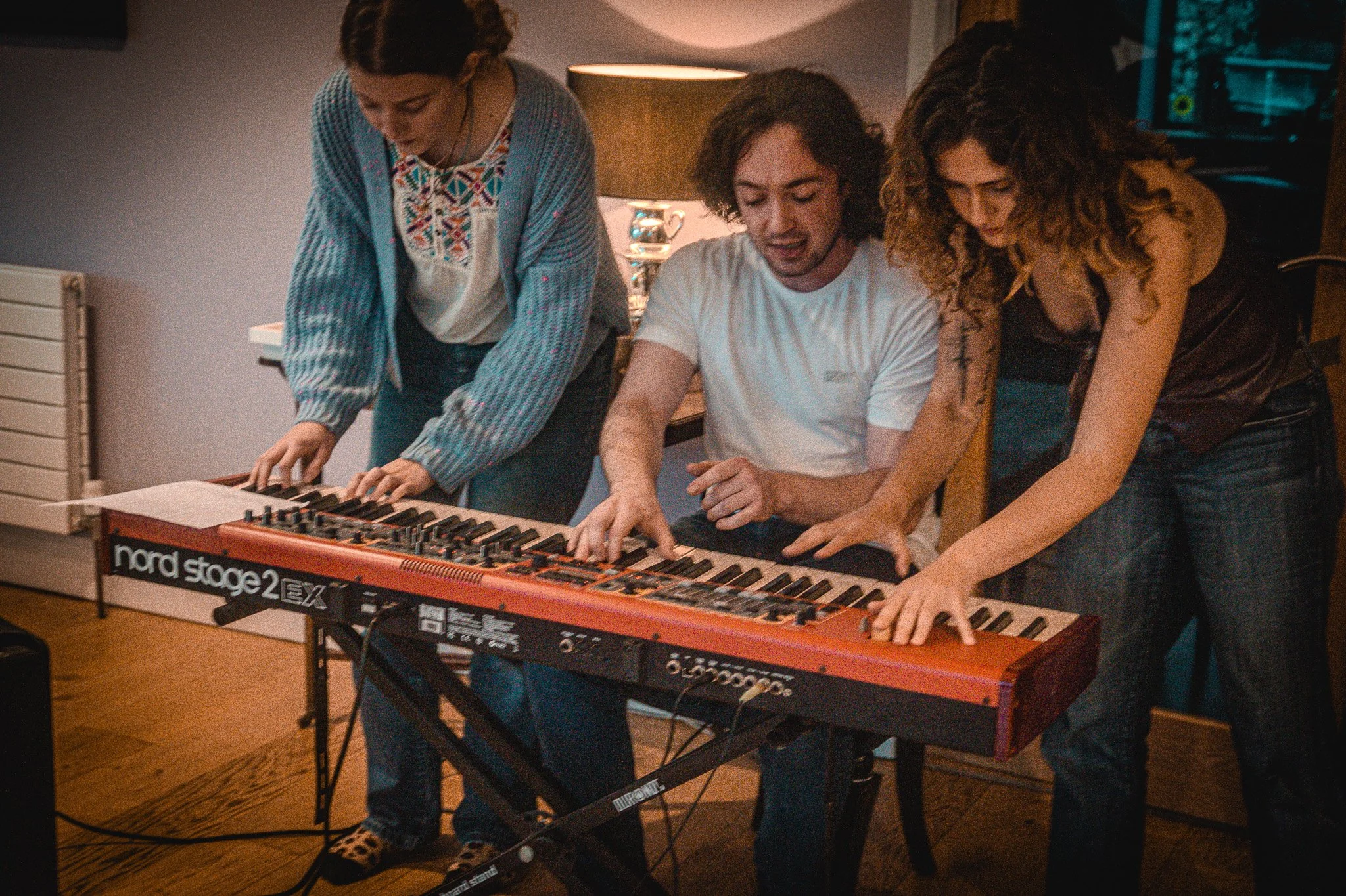 Three people gathered around a red Nord Stage 2 keyboard, with one person in a white t-shirt playing it, and the others leaning in to help or observe.