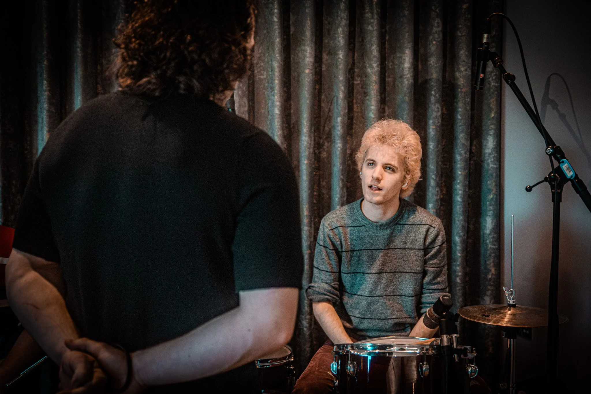 A person with light hair and a gray striped sweater sitting at a drum set, talking to a person with dark curly hair. They are indoors with dark curtains in the background and a microphone on a stand nearby.