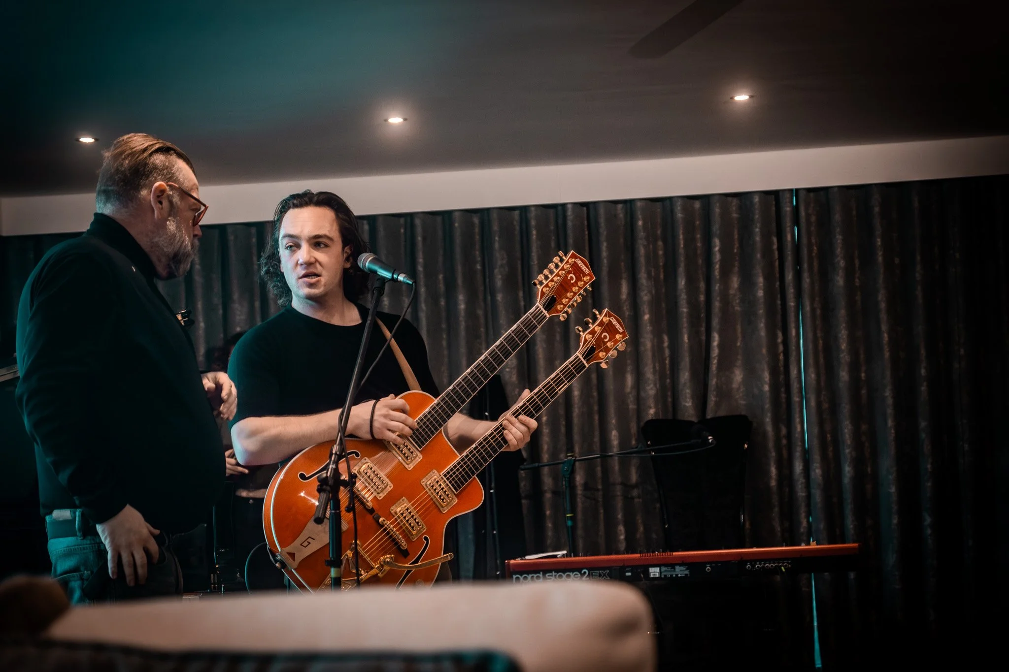 Two men on stage, one with a guitar, conversing during a performance in front of a dark curtain.