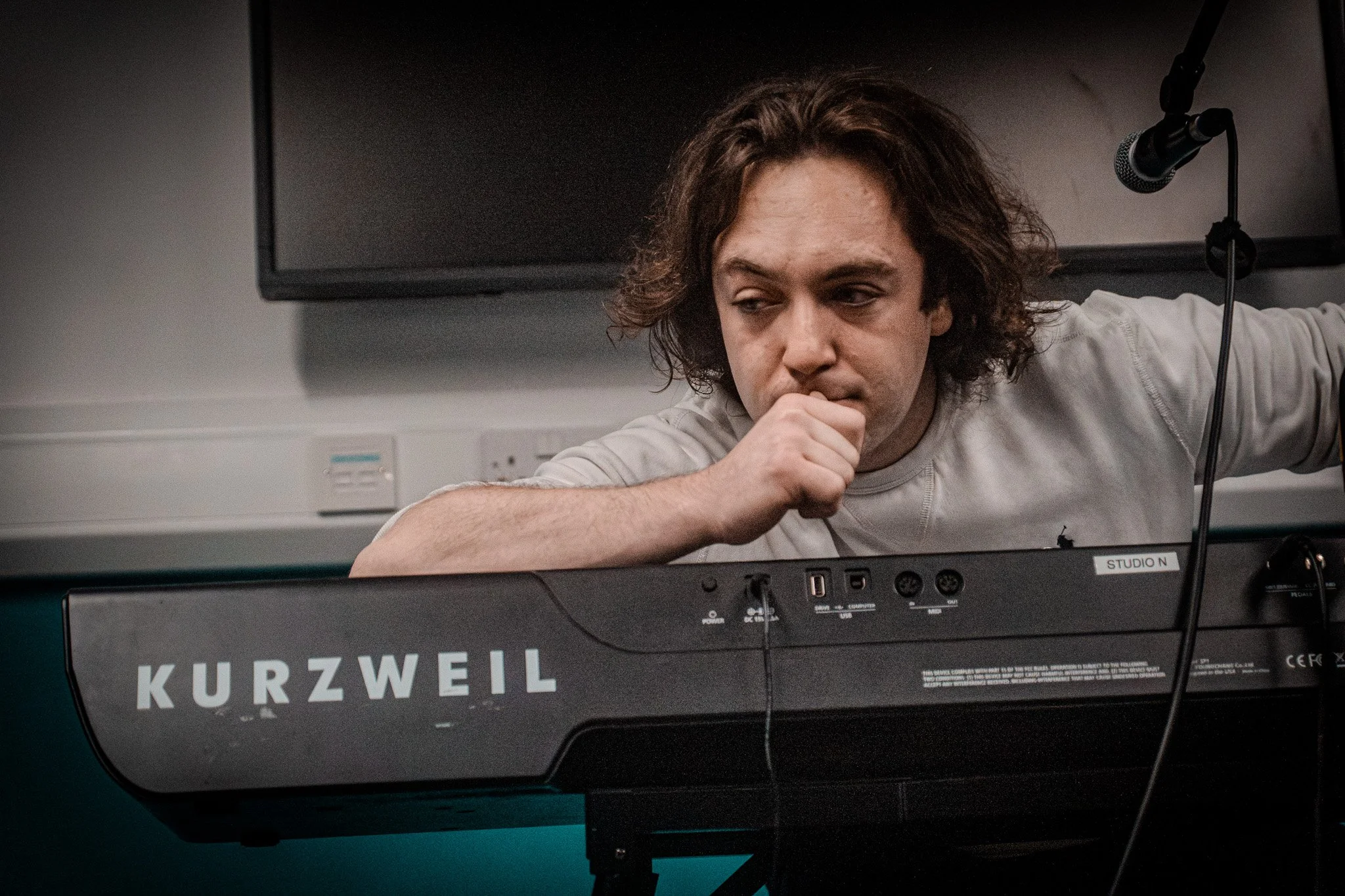 A man with long, curly hair resting his head on his hand, sitting behind a Kurzweil keyboard in a recording studio. There is a microphone in front of him and a large monitor screen behind him.