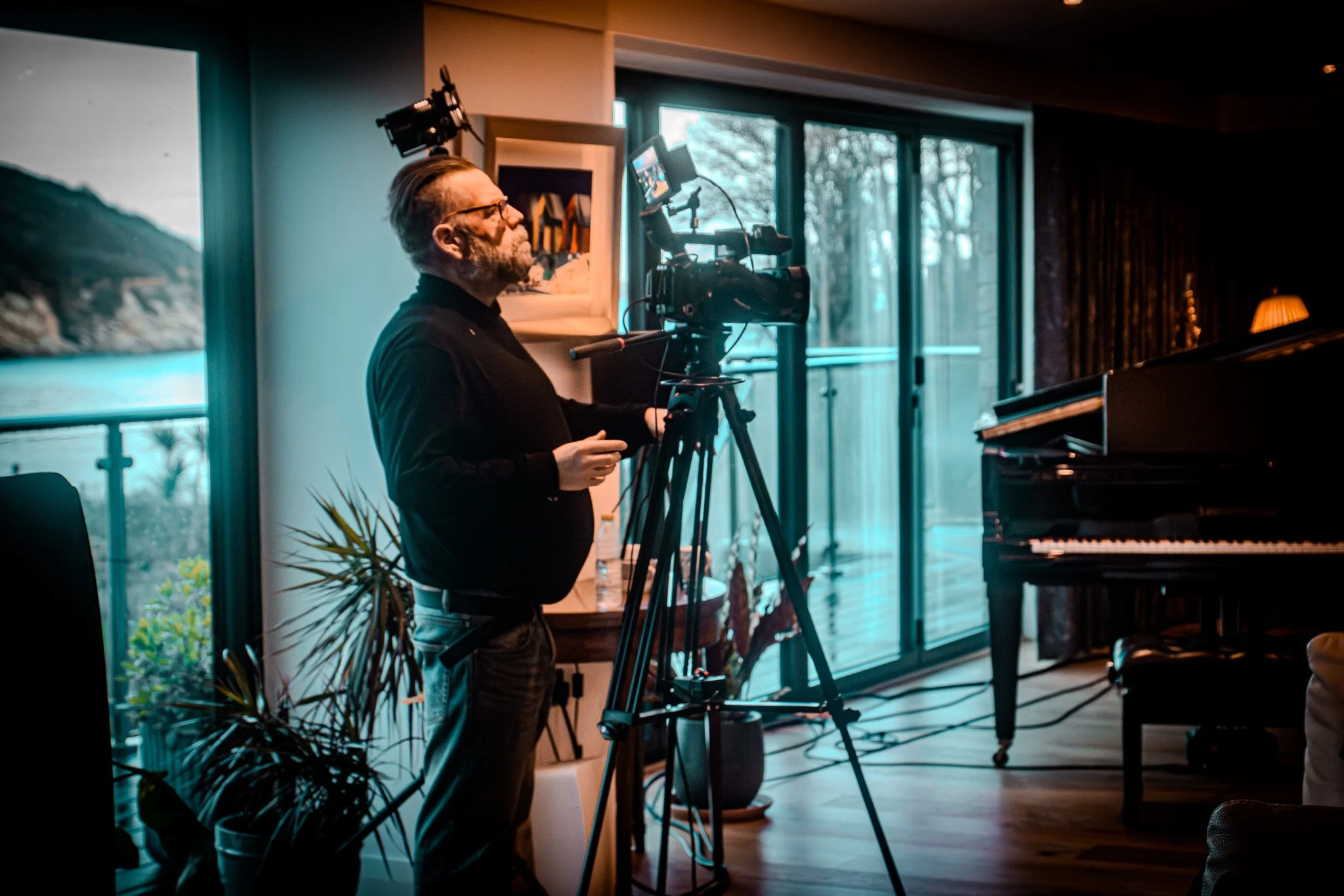 A man with glasses and a beard operating a professional video camera on a tripod indoors, near large glass doors with a view of trees outside. There is a piano visible to the right, potted plants, and artwork on the wall.