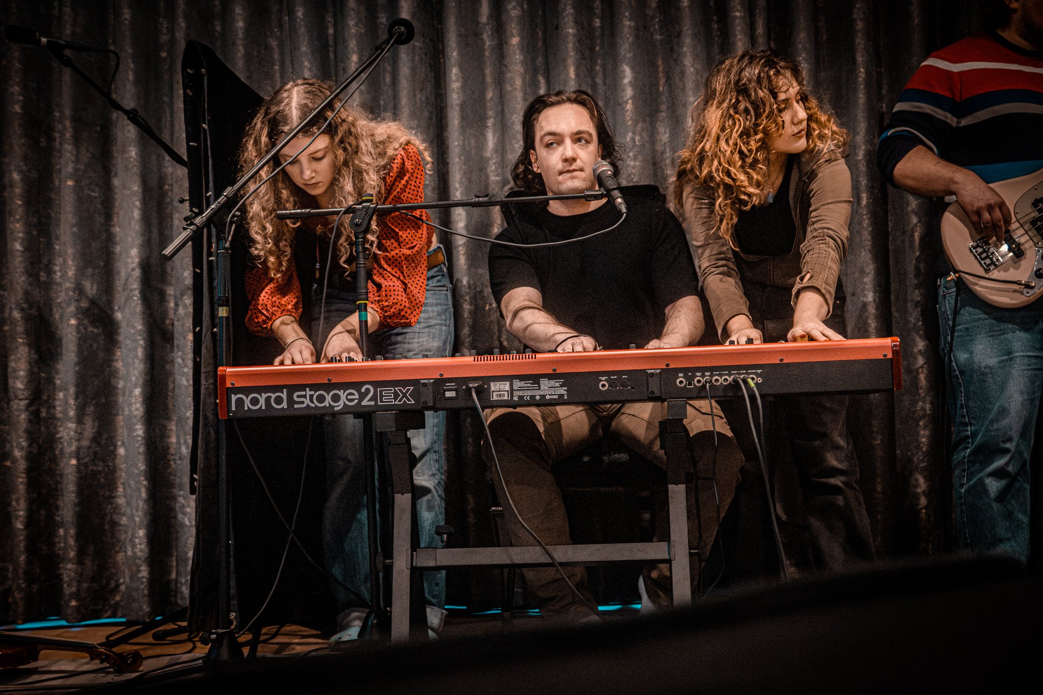 Three young women and a young man performing music on stage with a keyboard and microphone, with dark curtains in the background.