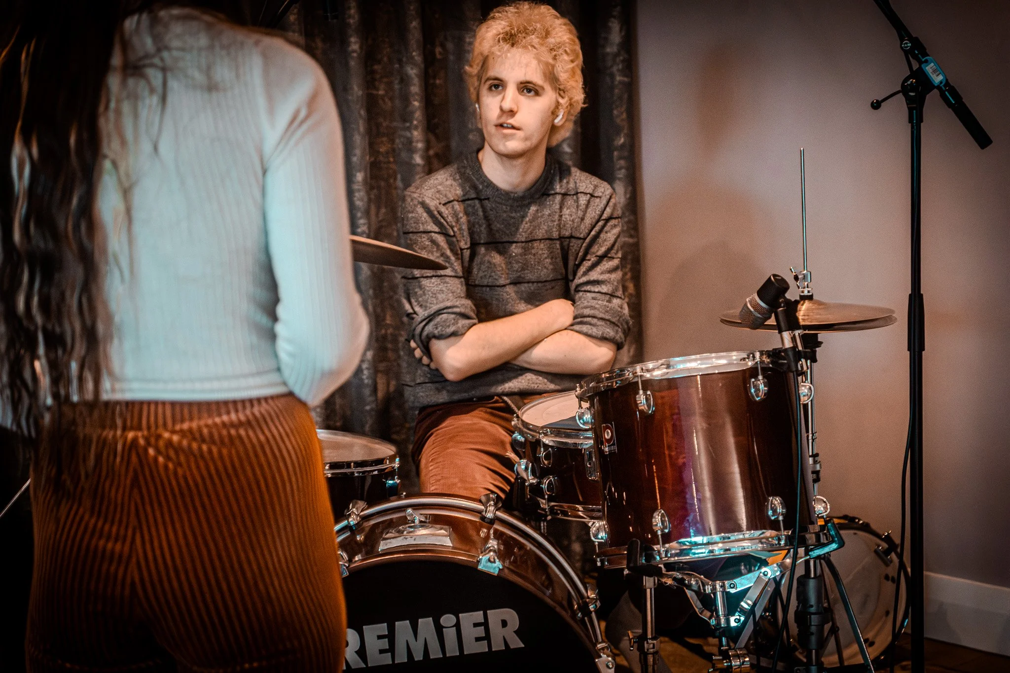 A young man with curly blonde hair wearing a gray and black striped sweater, sitting behind a drum set with arms crossed, appears to be listening or talking to a woman with long dark hair, white ribbed top, and brown corduroy pants in a music studio 