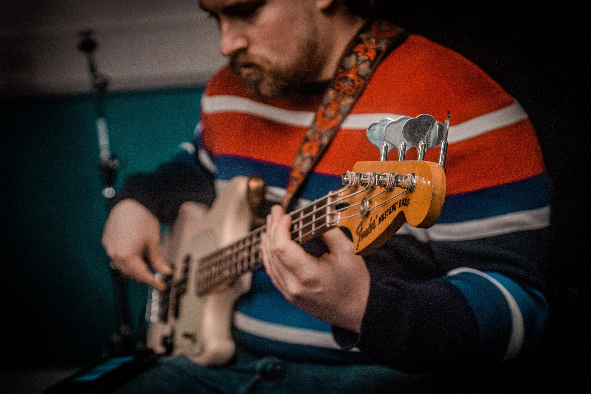 A man playing a ukulele in a room, wearing a multicolored striped sweater, with a patterned strap supporting the instrument, which has a label that reads 'Jamboree Mustangs'.