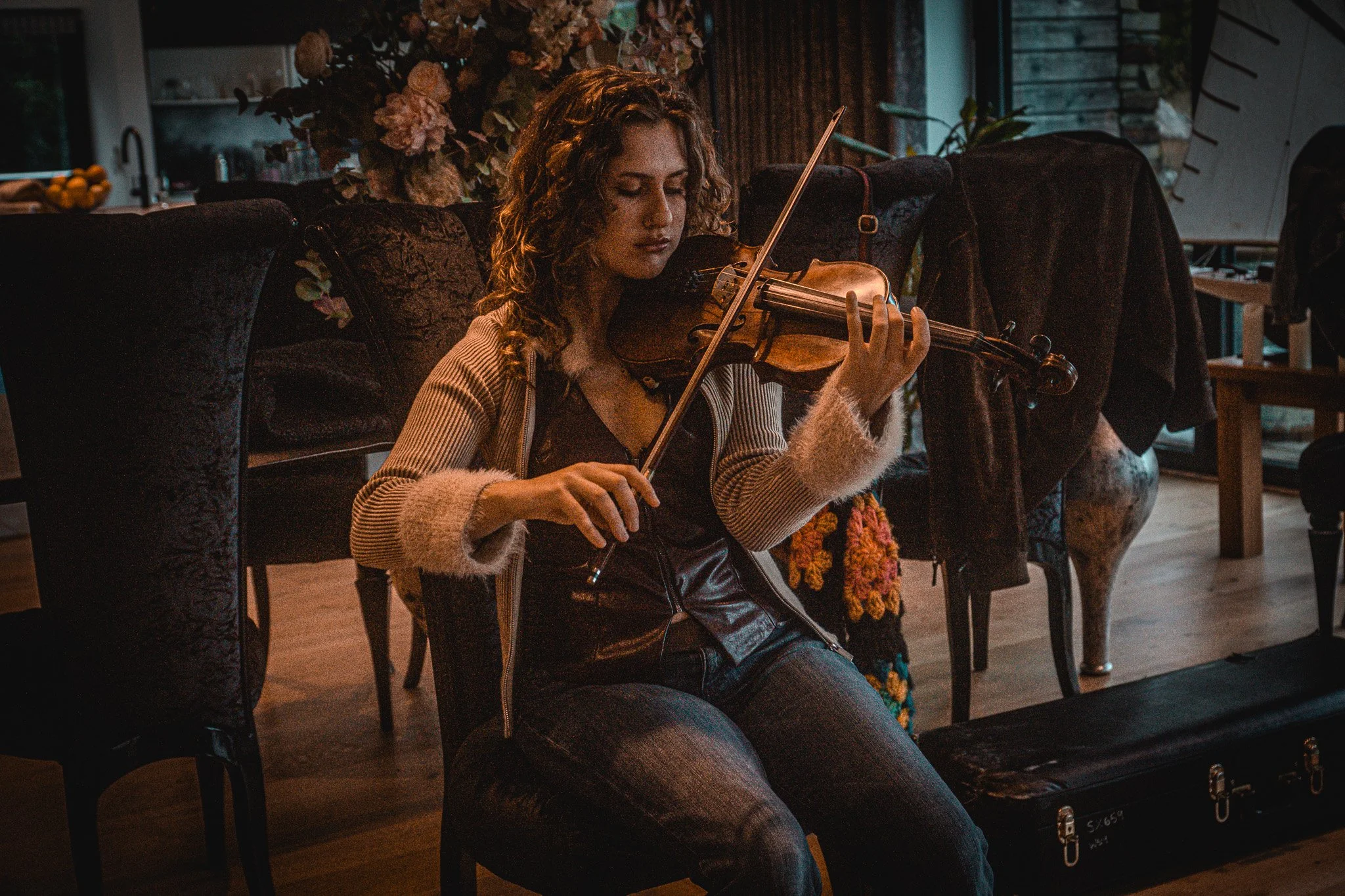 A woman playing the violin in a cozy, dimly lit indoor space, with flowers and furniture in the background.