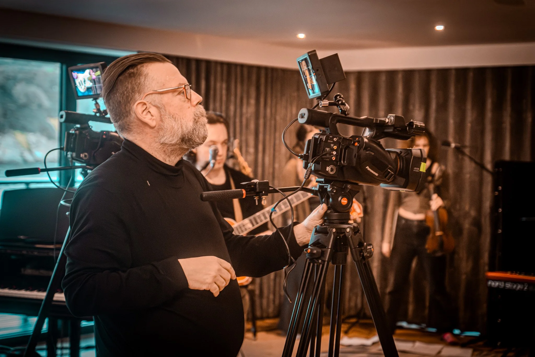 A man operating a professional video camera during a music recording session in a room with musicians in the background.