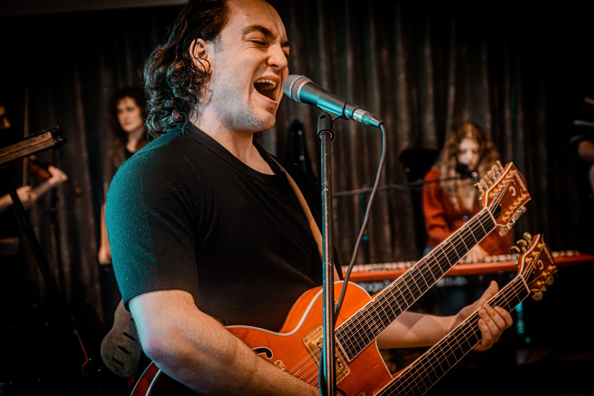 A man singing into a microphone while playing an orange double-neck guitar on stage, with other musicians in the background.