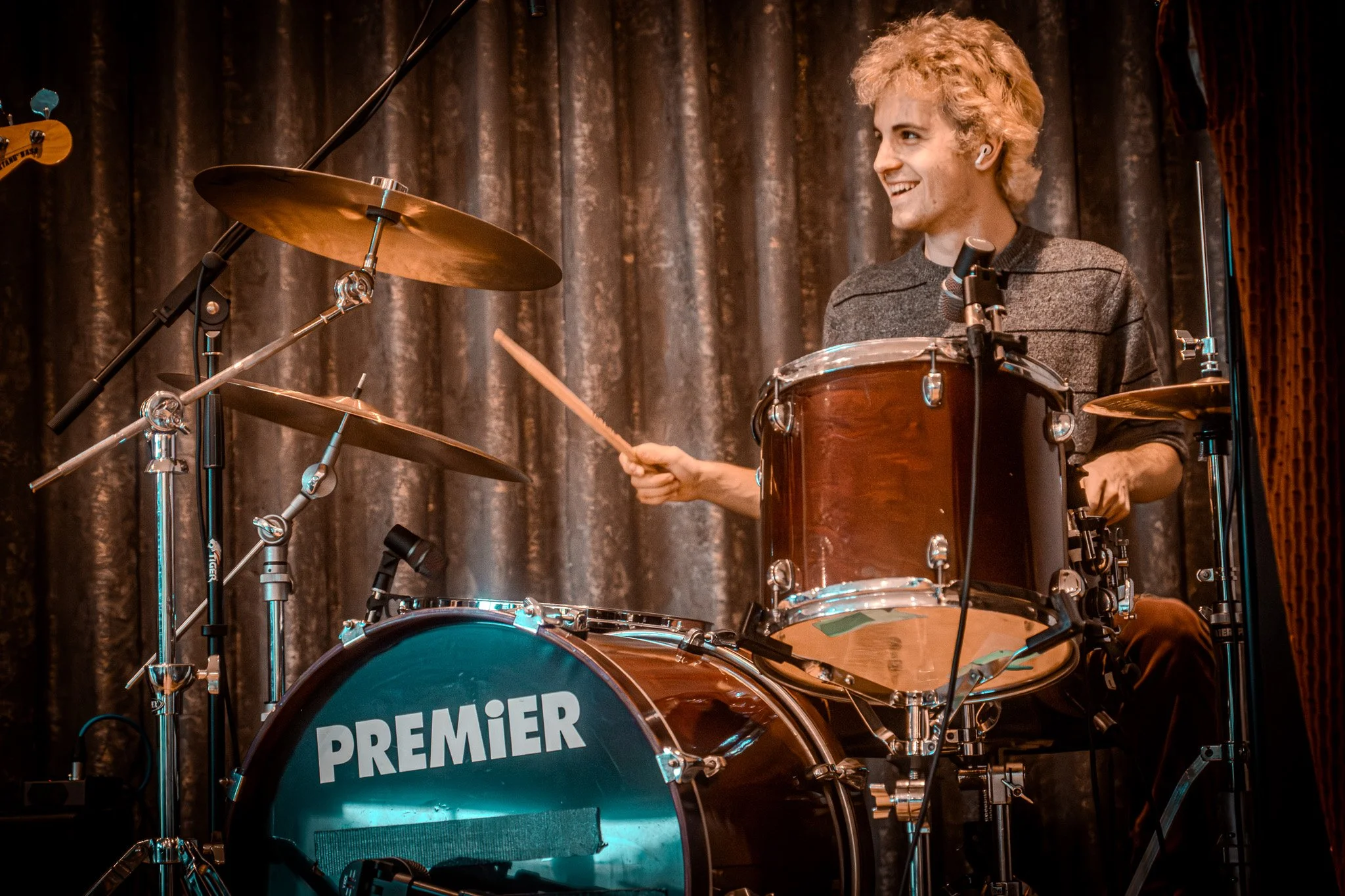 A young man with curly blond hair playing a drum set on stage, smiling, with a dark curtain in the background.