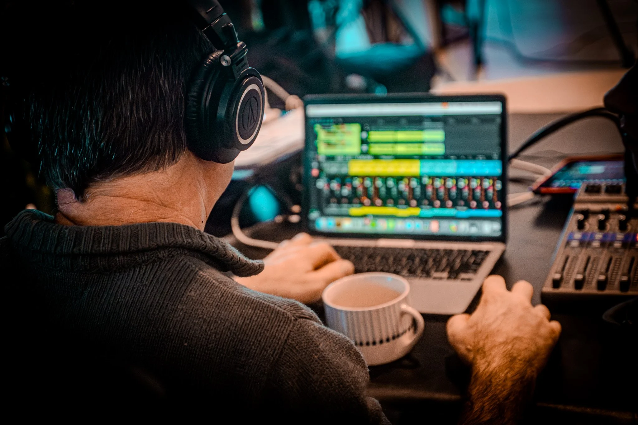 A person wearing large headphones working on a laptop with a digital audio workstation open, surrounded by audio equipment on a desk, with a mug nearby.