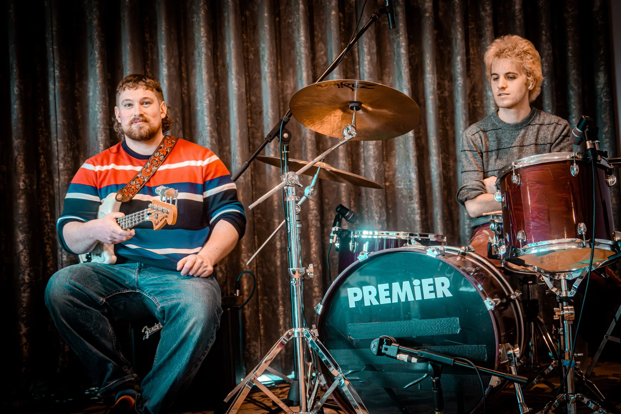 Two musicians on stage, one with a guitar sitting on a small amplifier, wearing a striped sweater, and the other with a drum set labeled "Premier," wearing a gray sweater, against a dark curtain backdrop.