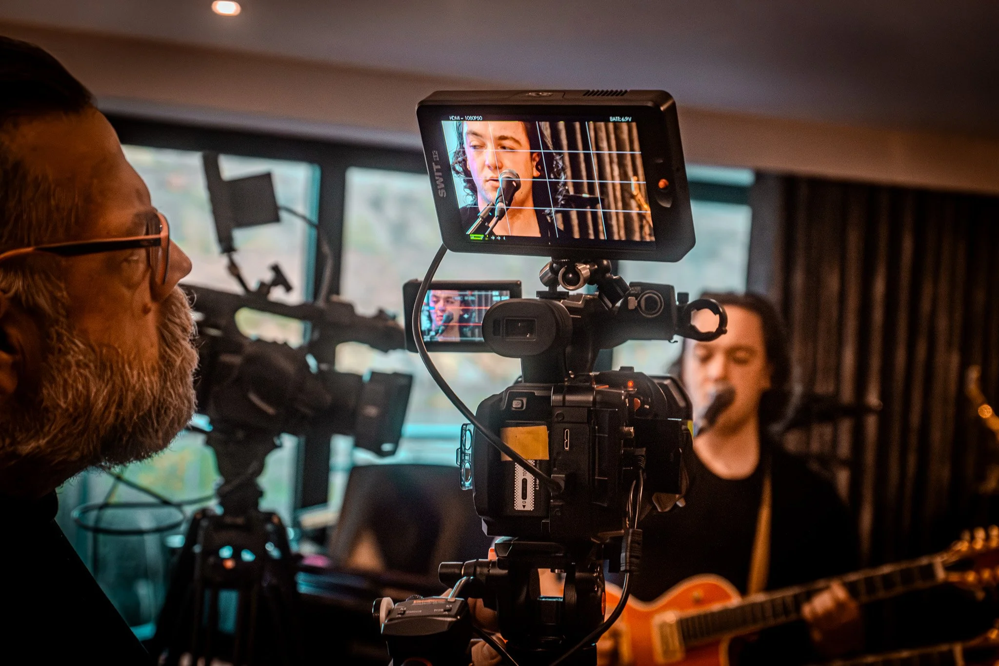 A man with glasses and a beard is filming a music performance with a professional video camera. The camera is focused on a musician playing guitar and singing into a microphone.