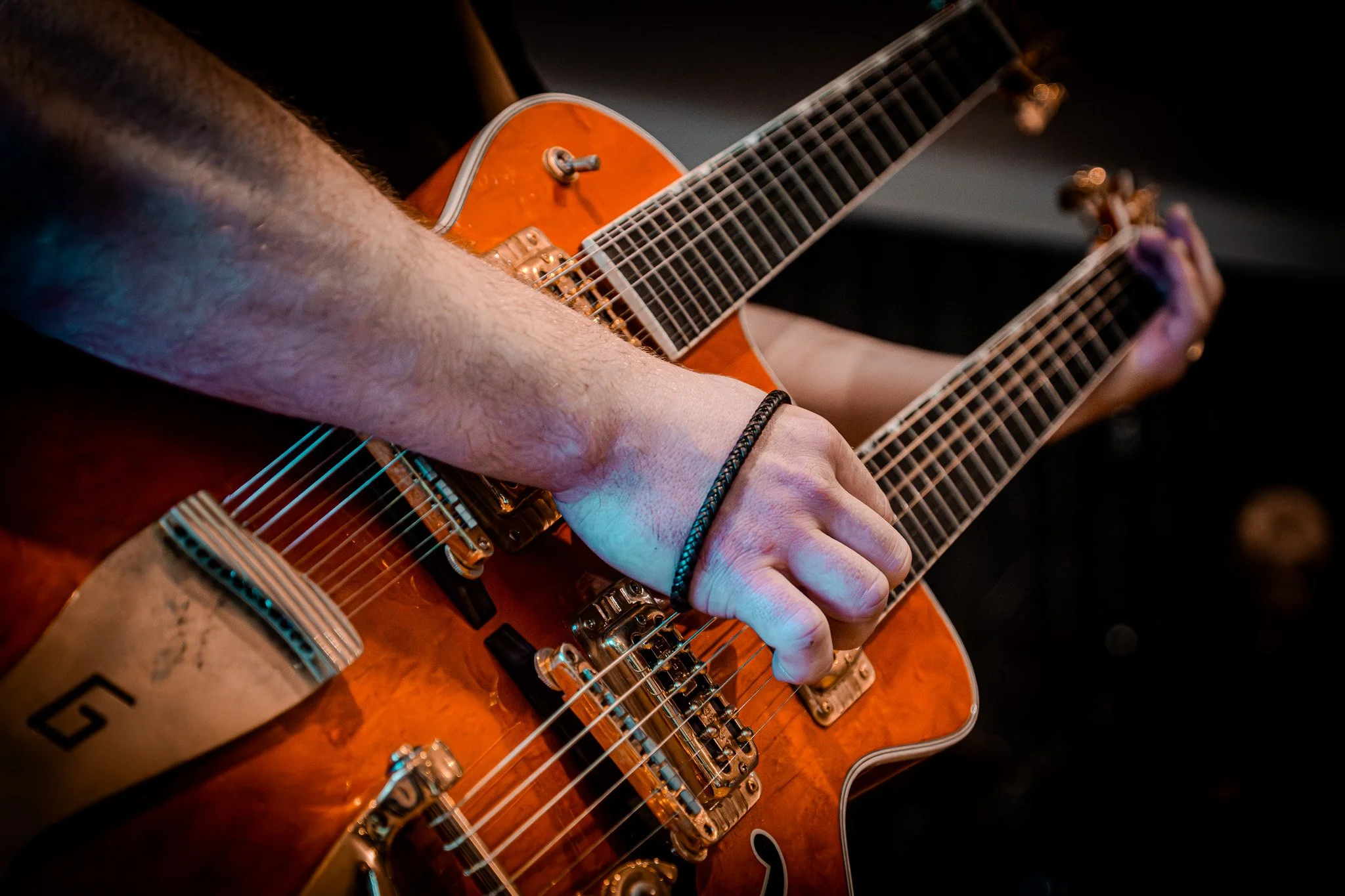 Close-up of a person's hands playing an orange bass guitar, with focus on fingers pressing strings and tuning pegs.