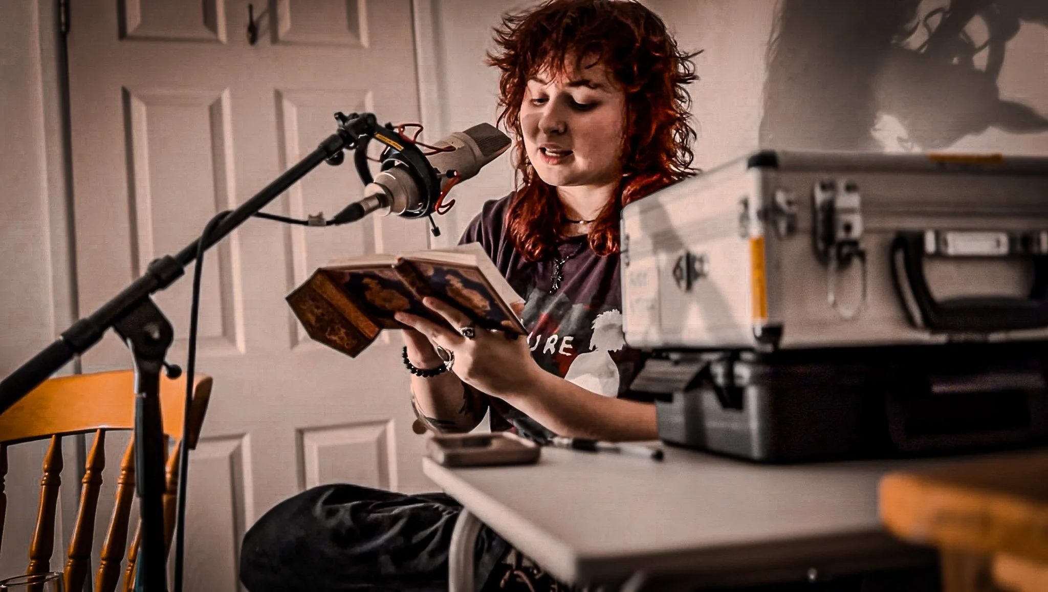 A woman with red curly hair sitting at a table, reading from a book and speaking into a microphone.