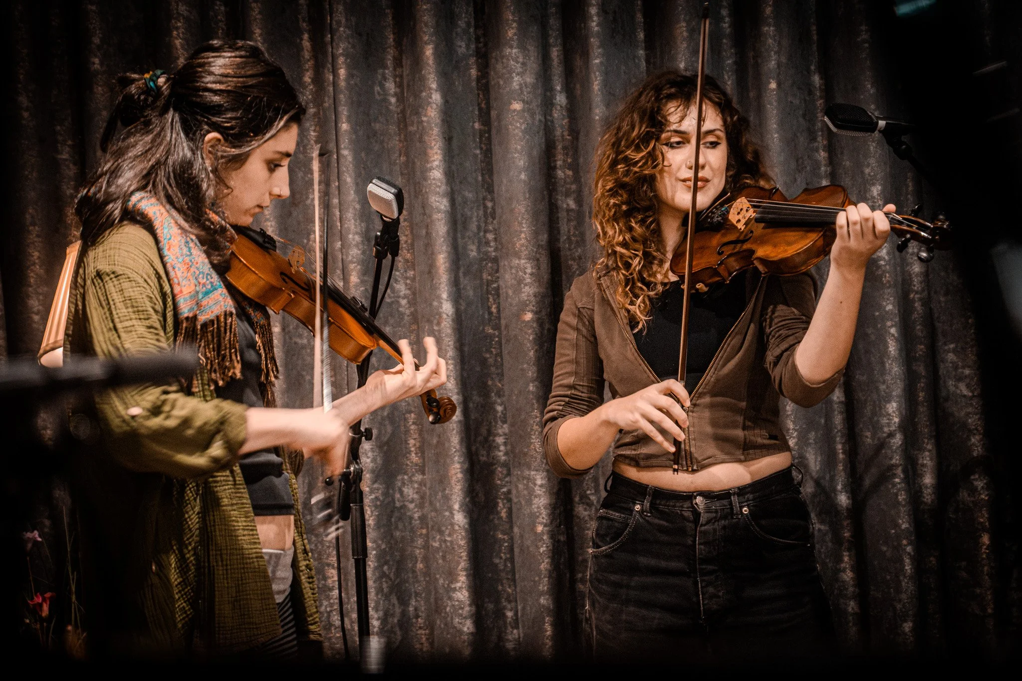 Two women playing violins on stage with a dark curtain backdrop.